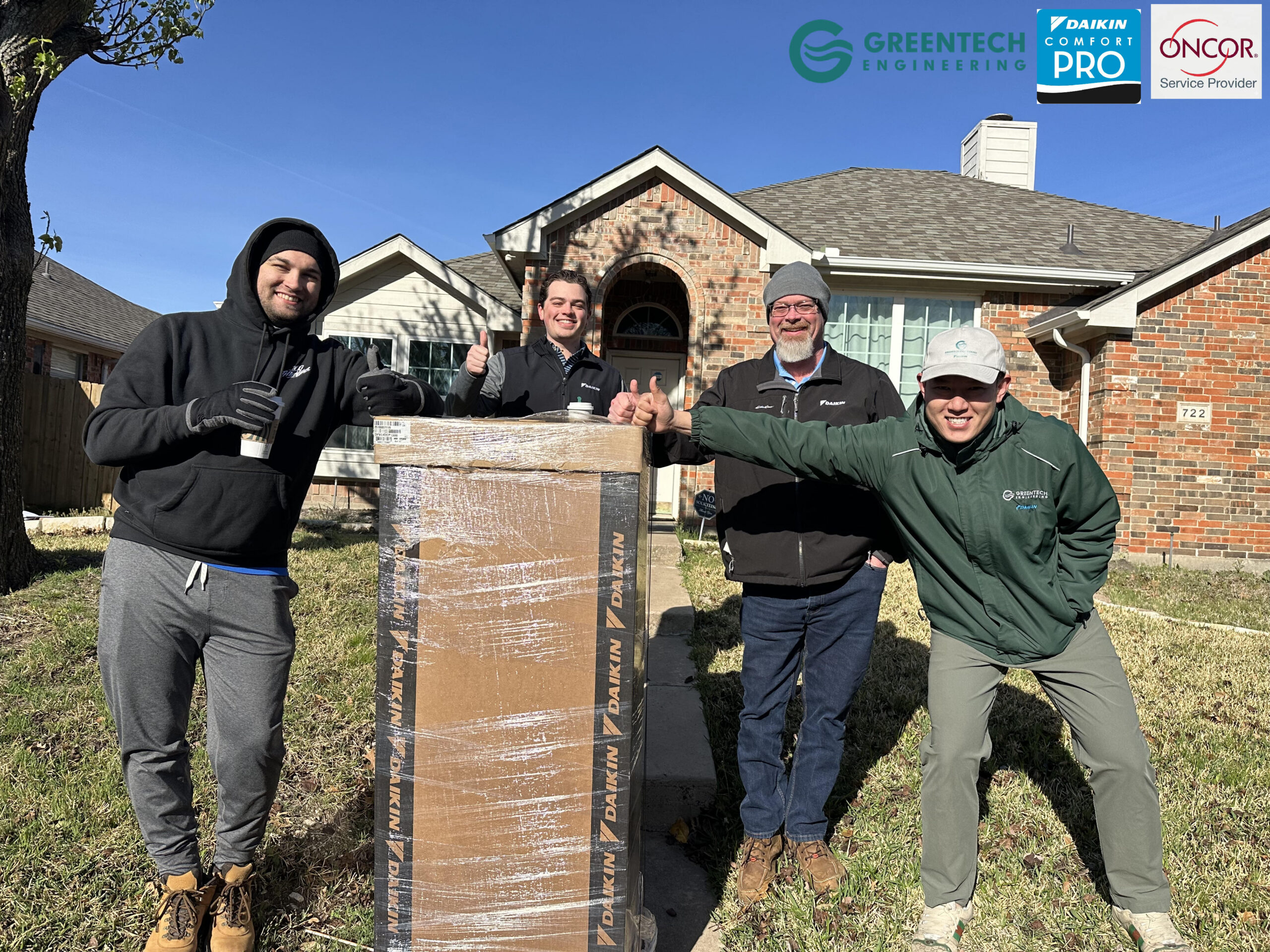 Greentech Engineering technicians smiling and giving thumbs up with a large packaged Daikin HVAC box on a residential lawn.