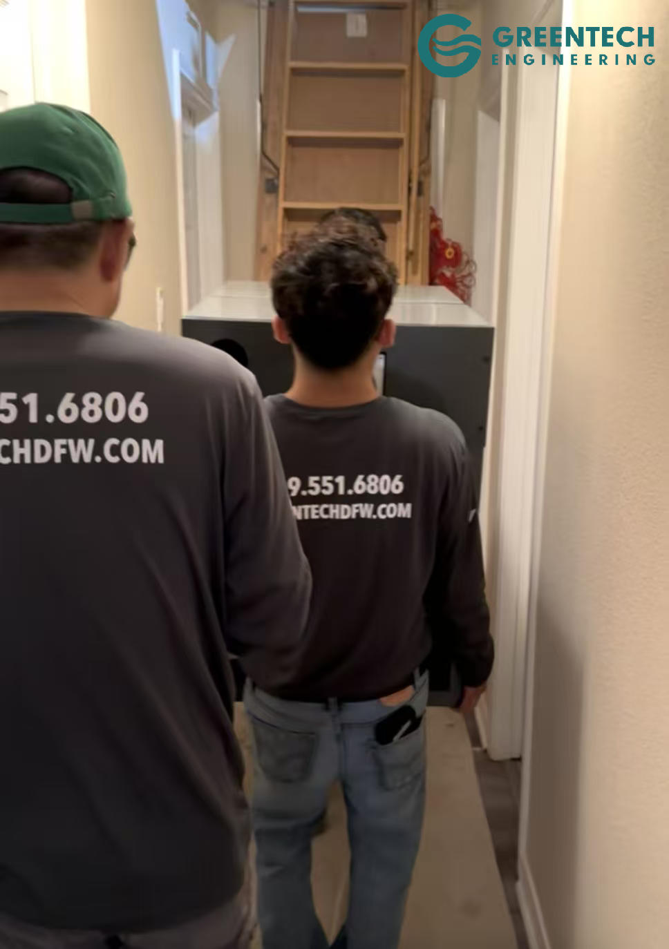 Two Greentech Engineering technicians carrying a high-efficiency Daikin air handler unit down a hallway to hoist it into the attic of a Dallas home.