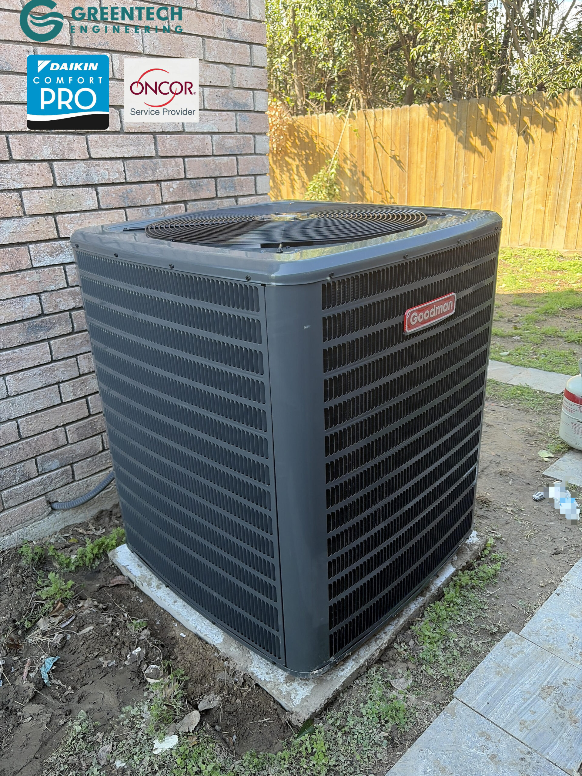 Completed installation of the grey Goodman outdoor condenser unit sitting on a concrete pad near a brick house.