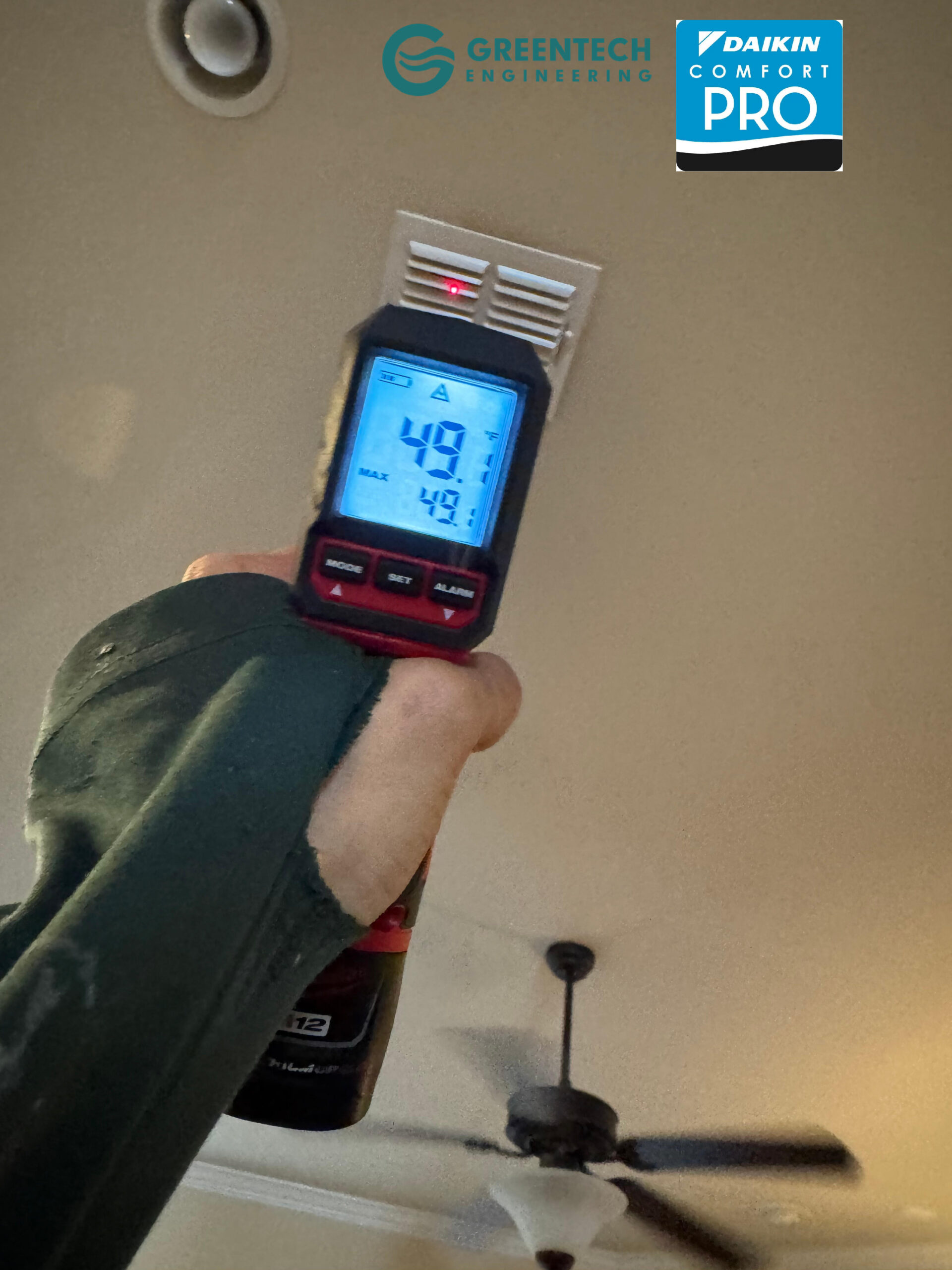 A technician using a digital infrared thermometer to measure a 49-degree supply air temperature coming out of a ceiling vent after a new Goodman AC installation.
