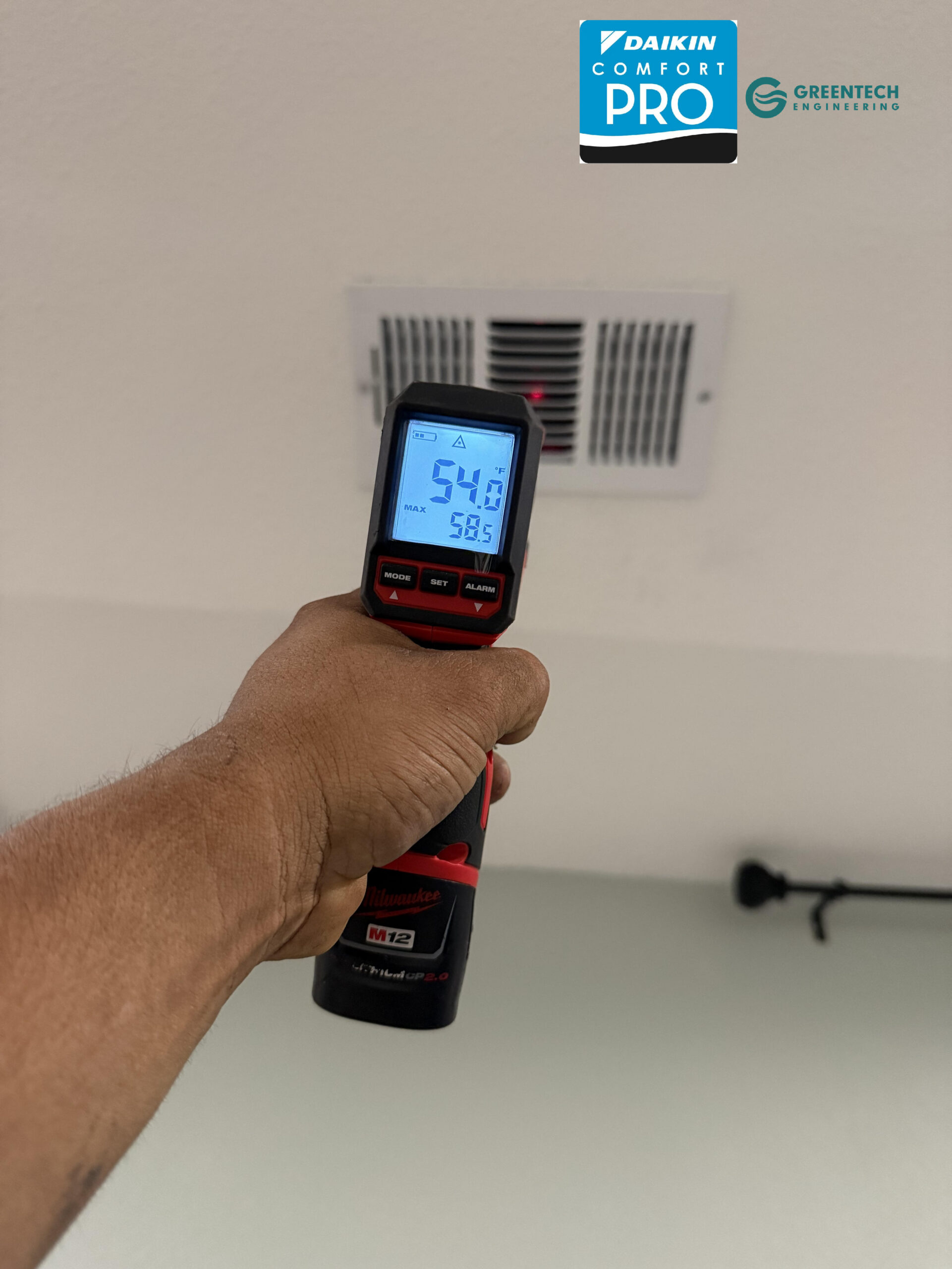 A technician's hand holding a Milwaukee digital infrared thermometer pointing at a ceiling vent, reading a supply air temperature of 54.0 degrees Fahrenheit.