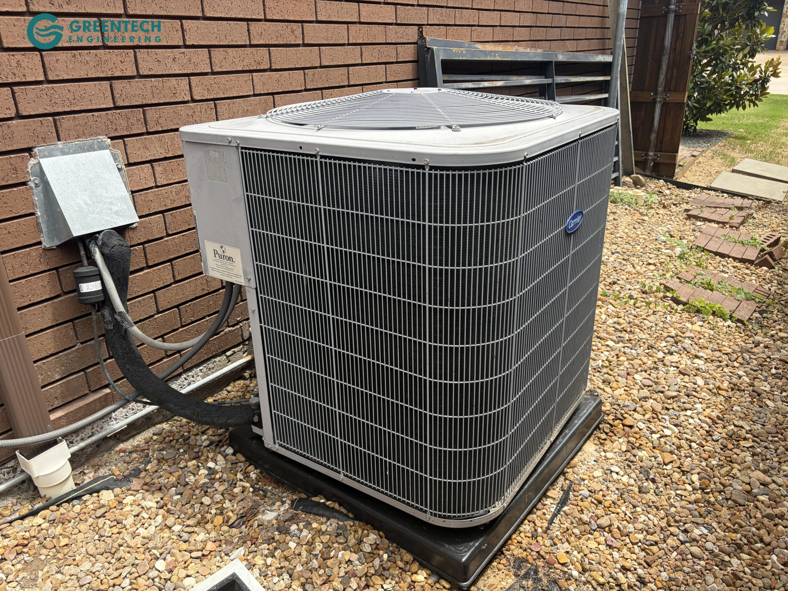 An aging, inefficient central air conditioning condenser sitting on a black plastic pad outside a brick two-story home in Dallas, Texas 75248.