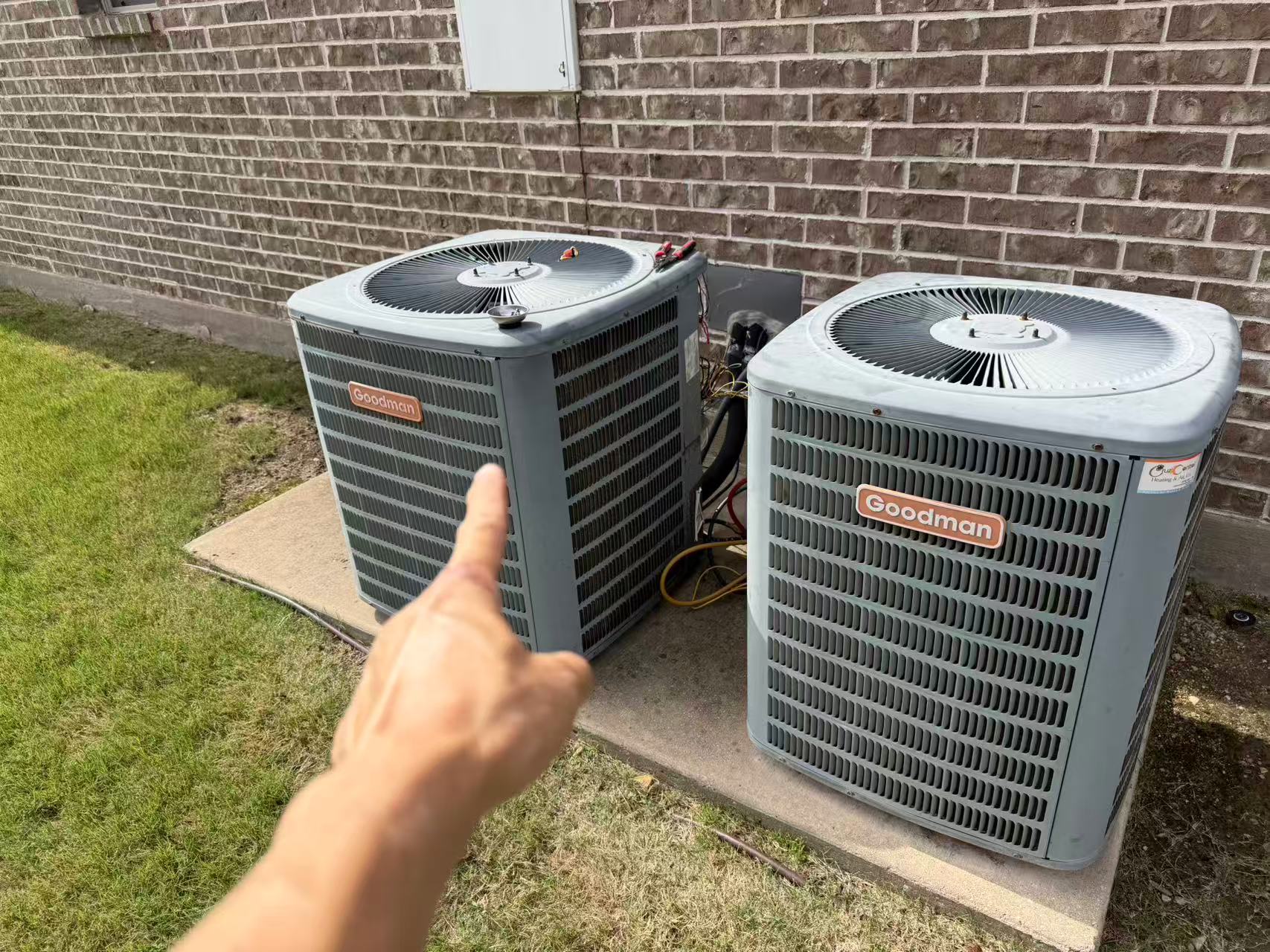 An HVAC technician pointing at an aging, inefficient Goodman air conditioning condenser sitting on a concrete pad in a Dallas backyard.