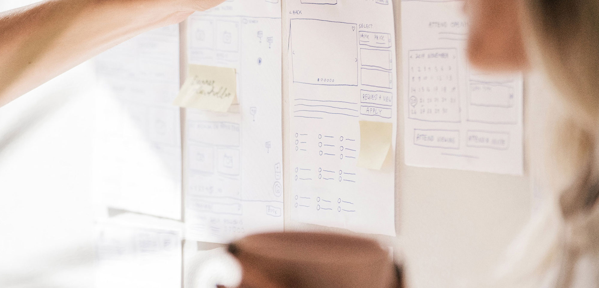 Close-up of hand placing sticky notes on wall with wireframe sketches and calendar design mockups.