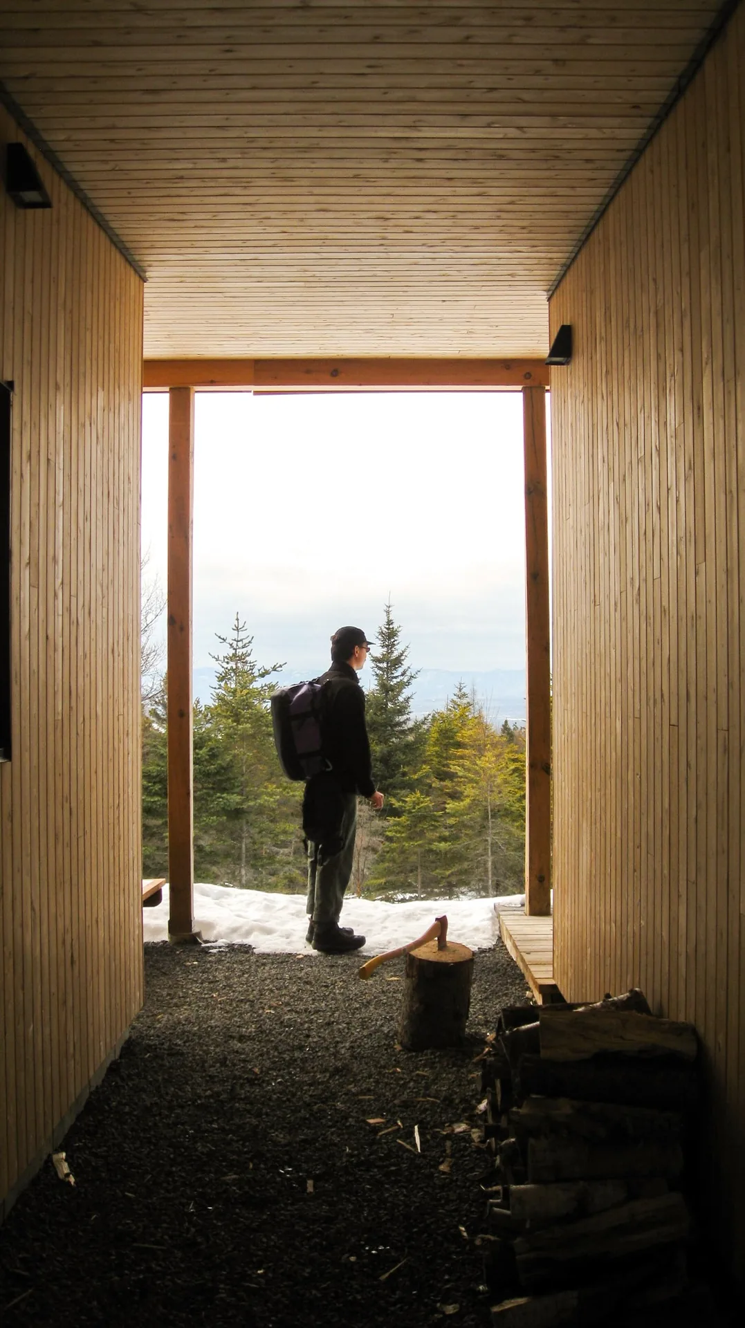 Vue depuis l'intérieur d'un refuge en bois vers un randonneur avec sac à dos devant une forêt enneigée dans Charlevoix