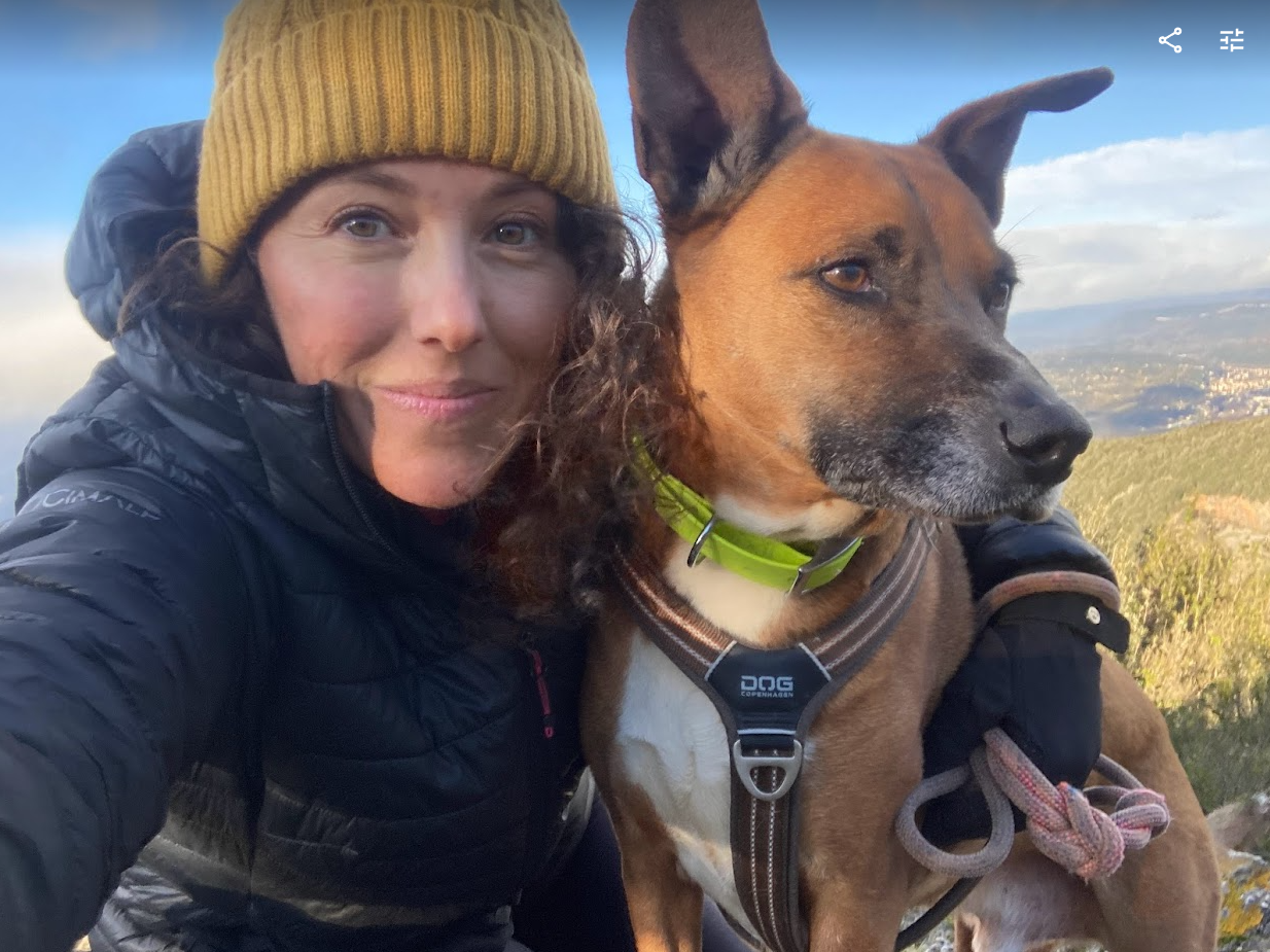 Marine Coach Canin porte un bonnet et pose avec son superbe chien roumain Daima, avec un magnifique paysage du Parc Naturel Régional du Haut Languedoc. Ils ont le visage serein.