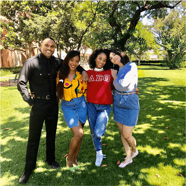 Behind-the-scenes shot of four young actors, one in a cop outfit and two women in denim skirts, standing together on a grassy lawn under trees, smiling. 