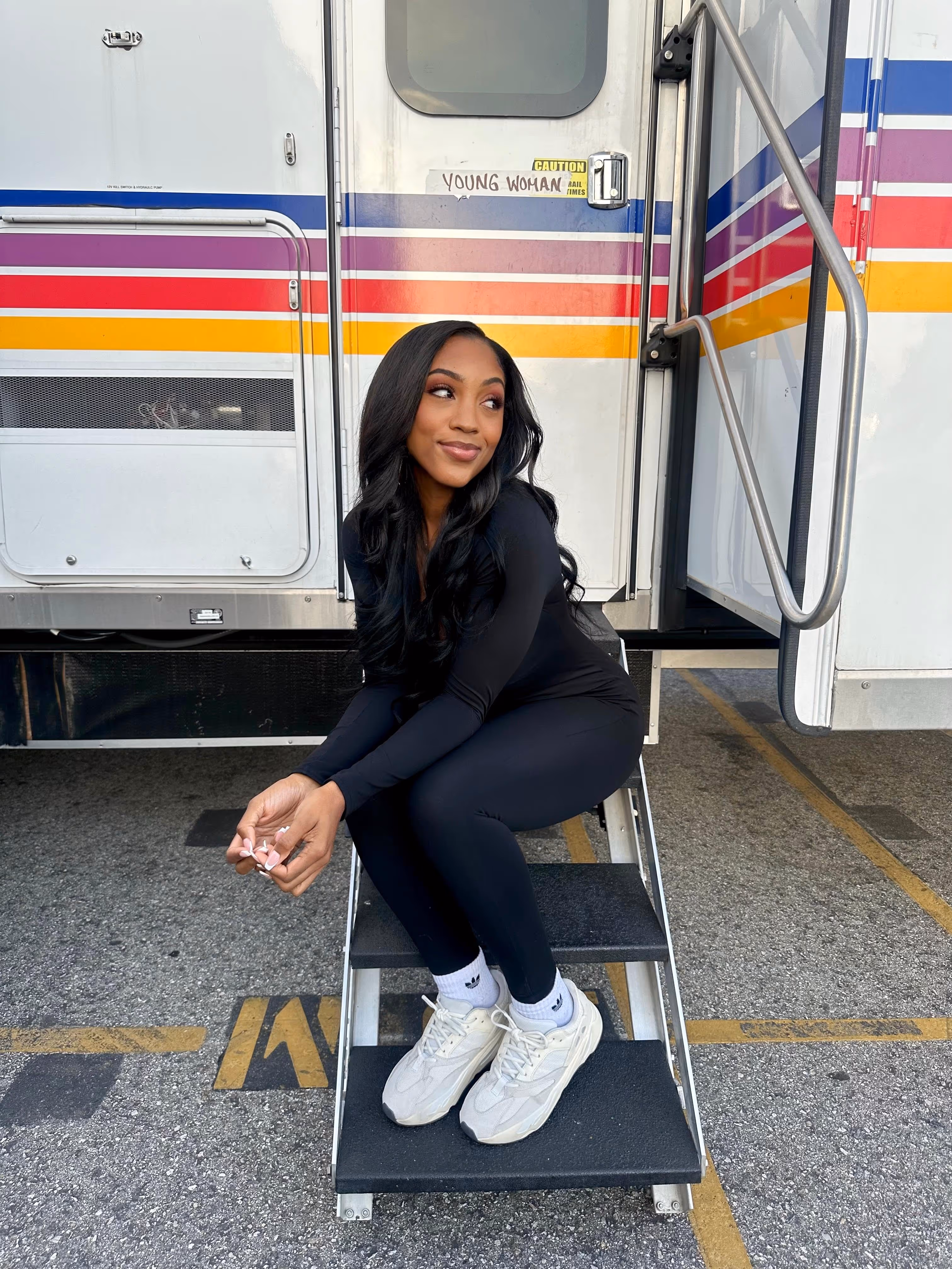 Actress  in comfortable black athleisure wear on trailer steps, with a 'YOUNG WOMAN' sign visible above the door. 