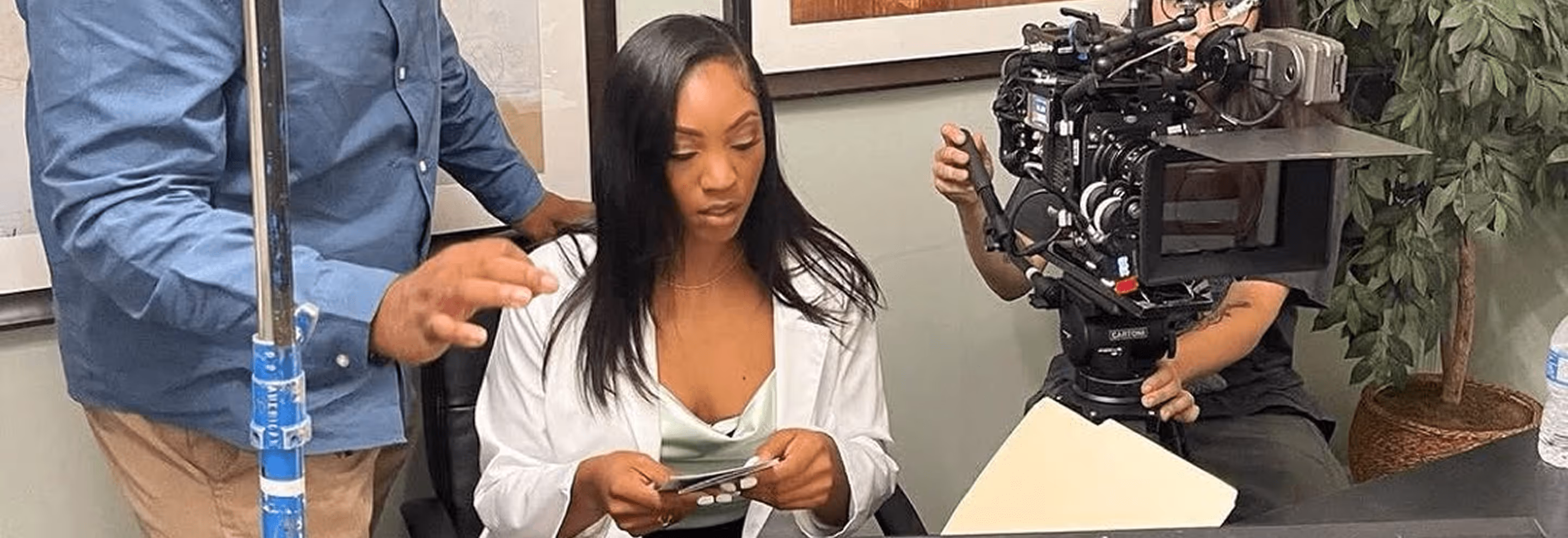 Behind-the-scenes shot of a film or photoshoot with a man holding a boom pole, woman going over a deck of index cards, and a professional video camera.