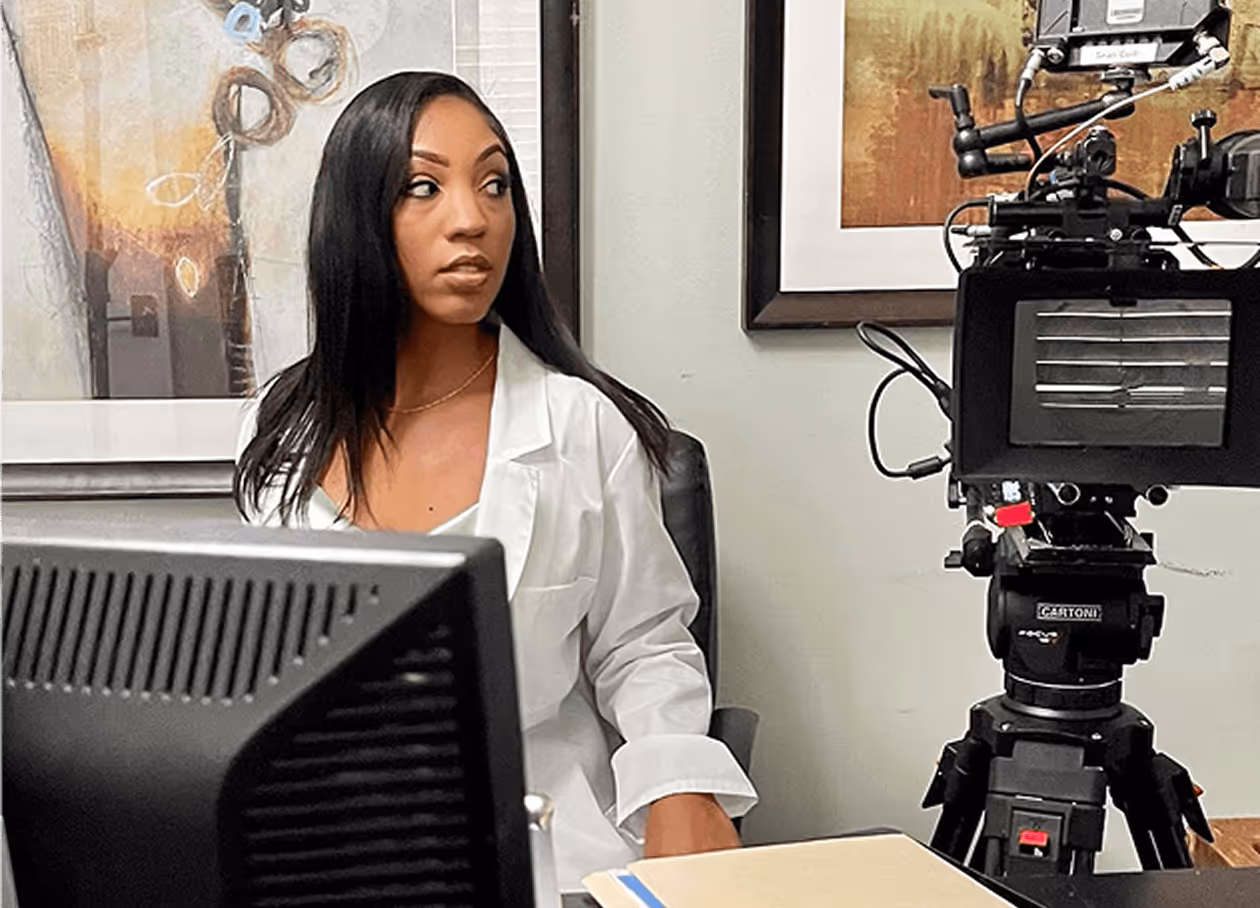 Actress in a white lab coat on a film set with professional camera & computer monitor.