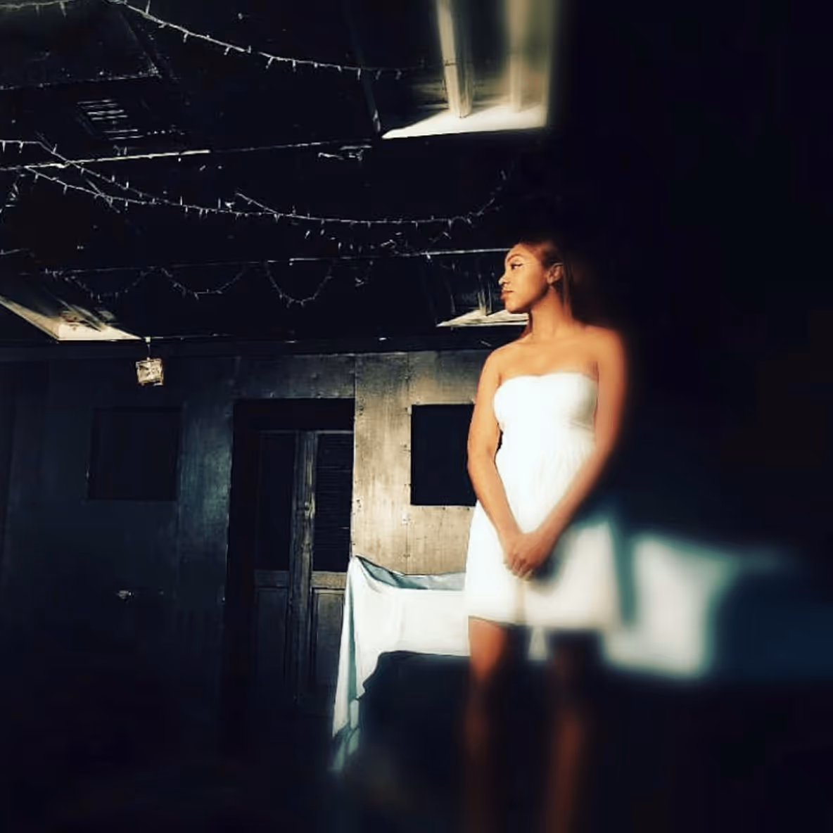 Actress on a dark, moody set, wearing a white dress and looking off to the side.