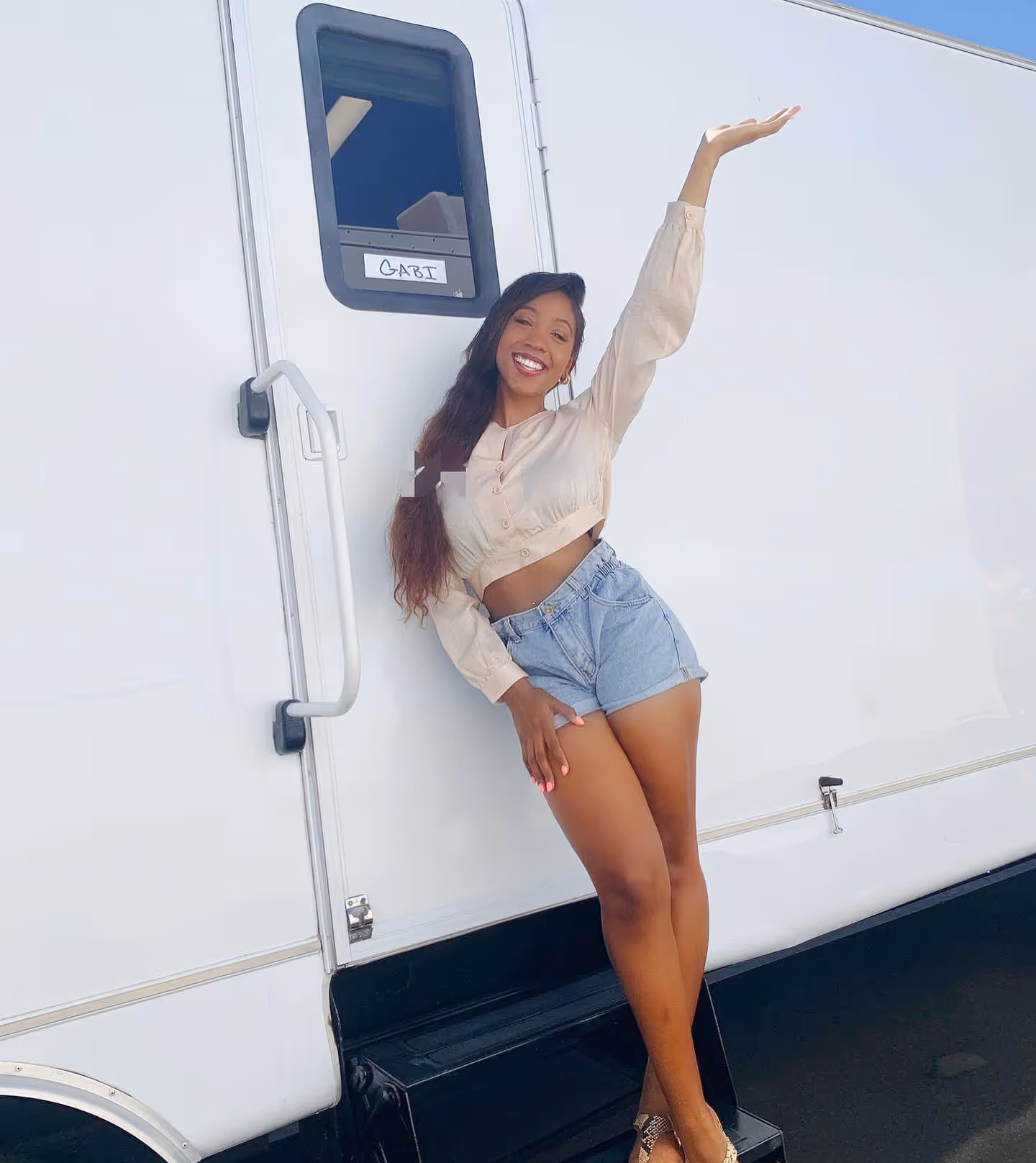 Smiling woman in a crop top and denim shorts posing playfully next to a white trailer door labeled 'GABI'.
