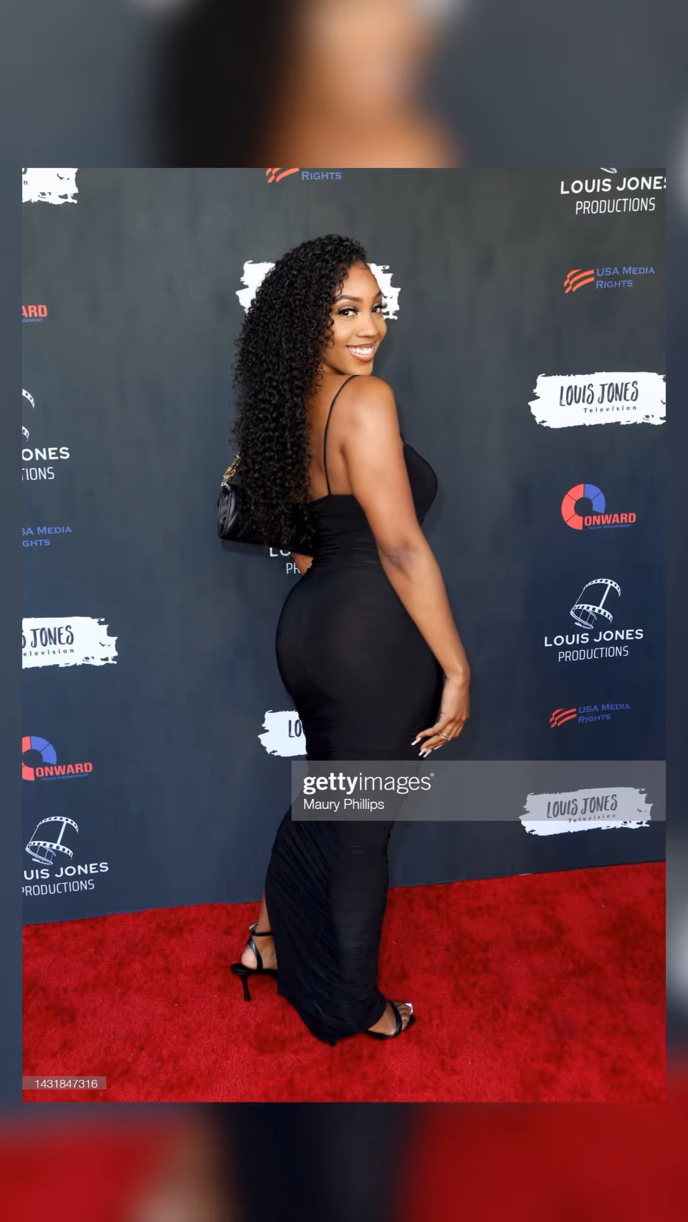 Woman in a fitted black dress and heels smiling over her shoulder on a red carpet, with a backdrop of sponsor logos including 'Louis Jones Productions'.