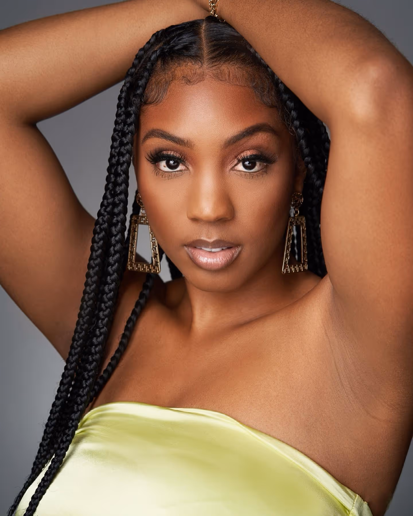 Portrait of a confident woman with braided hair and striking makeup, looking directly at the camera, against a neutral background. 