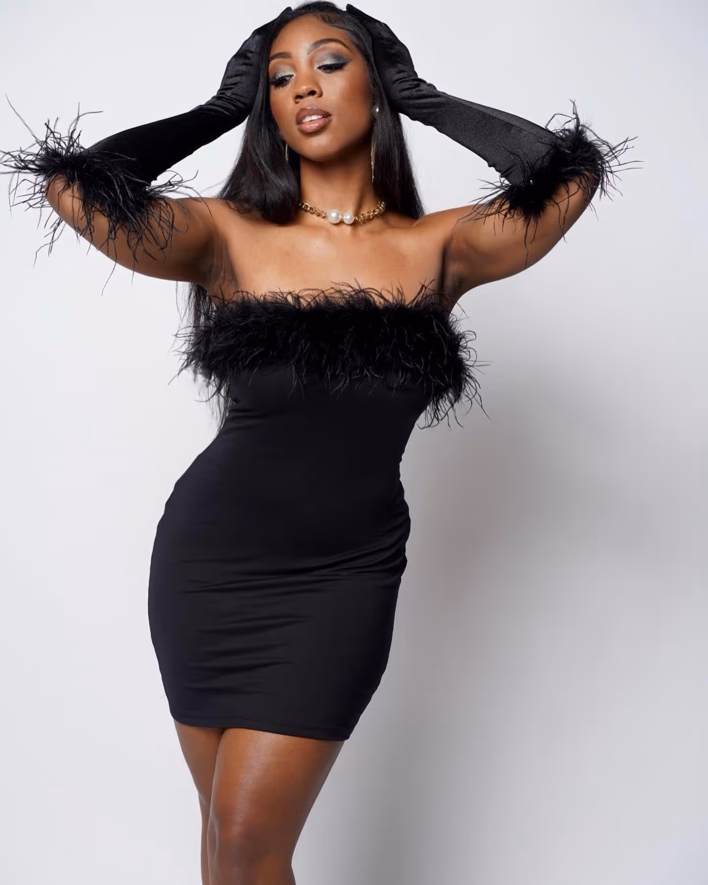 Glamorous woman in a strapless black feathered dress and long black gloves, posing elegantly with hands on her head. 
