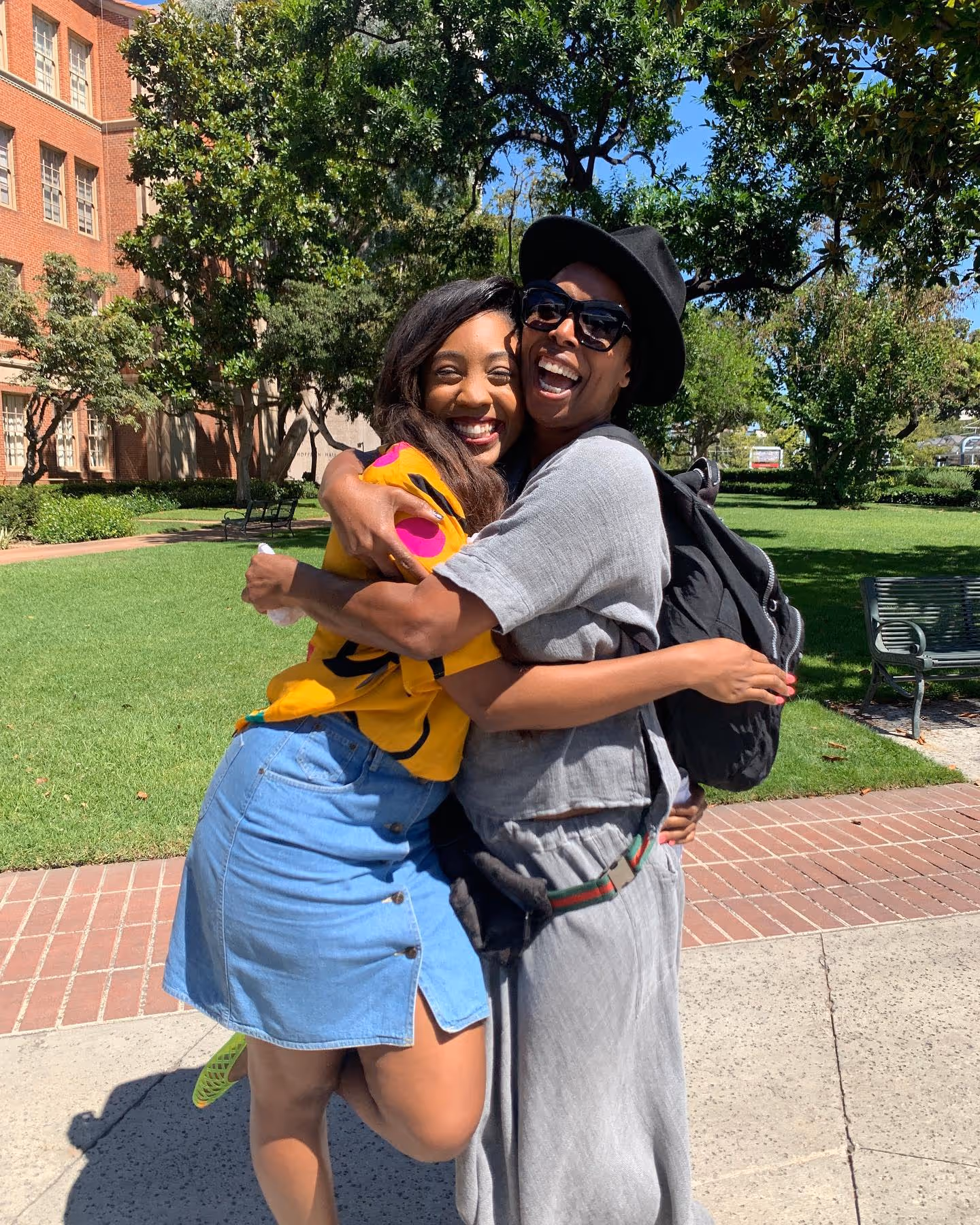 Two smiling women embracing outdoors on a sunny campus or park-like setting. 