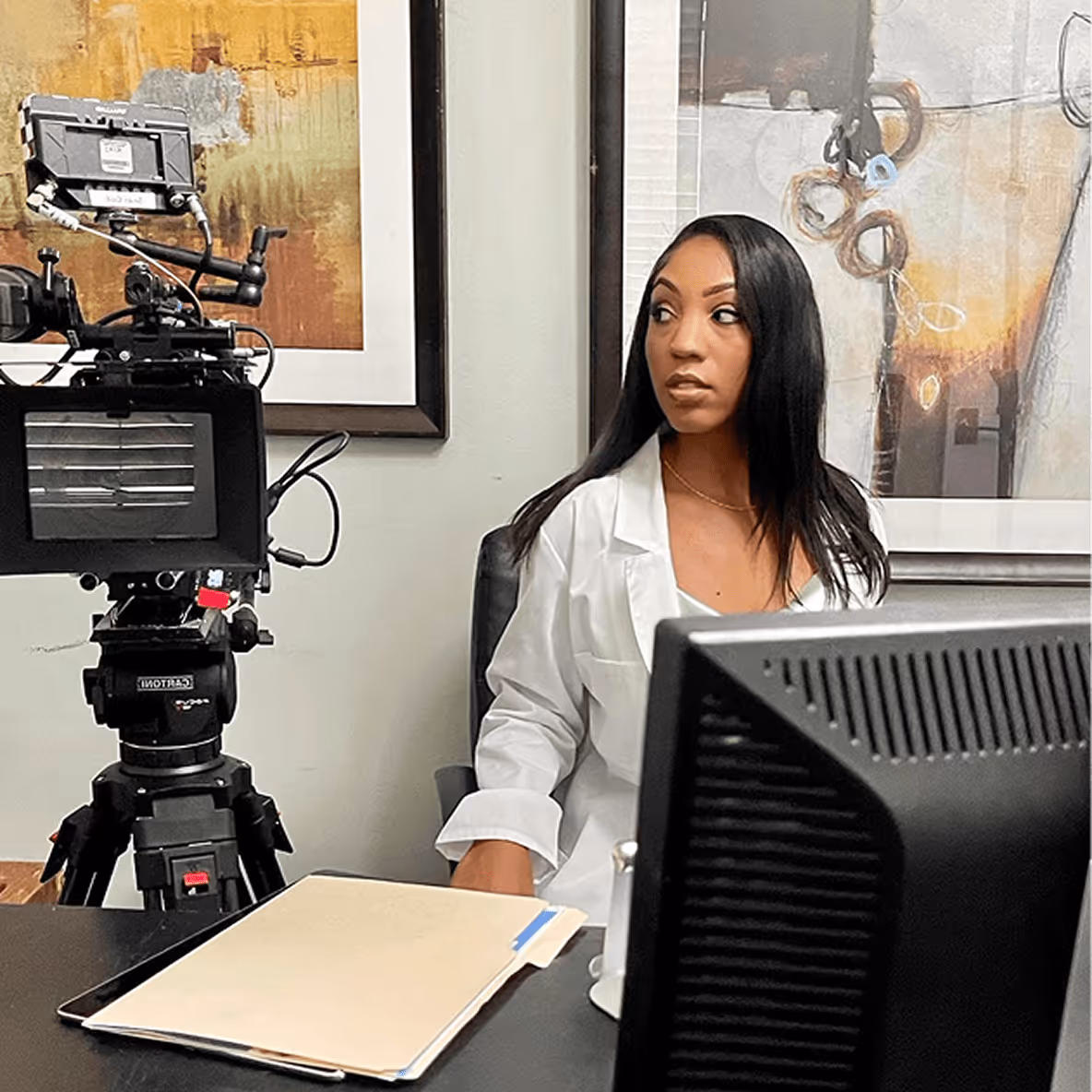 Woman in a white lab coat sitting at a desk with a monitor and film camera. 