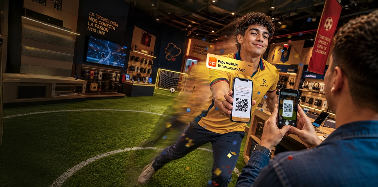 Vendedor cobrando a un cliente con código QR Payphone desde su celular en tienda de tecnología con temática de fútbol.