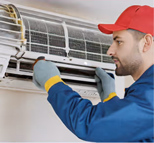 A technician in a blue uniform services an air conditioning unit indoors.