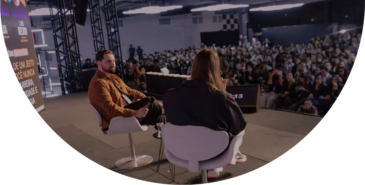 Duas pessoas sentadas em cadeiras em um palco, participando de uma conversa diante de uma grande plateia.