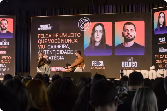 Palestra com Felca e Luan Beluco no palco do Influent Summit diante de uma plateia grande, com tela ao fundo mostrando fotos e nomes dos palestrantes.
