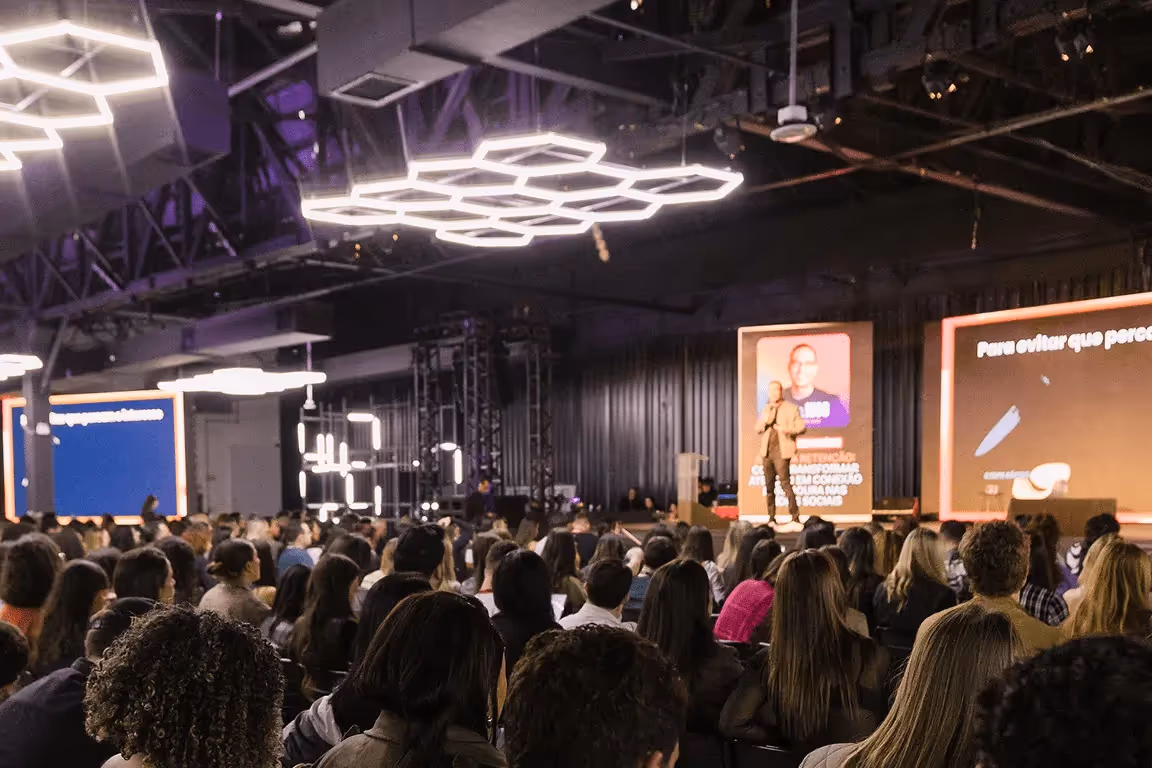 Auditório cheio com público assistindo palestra de homem em palco com telas iluminadas ao fundo.