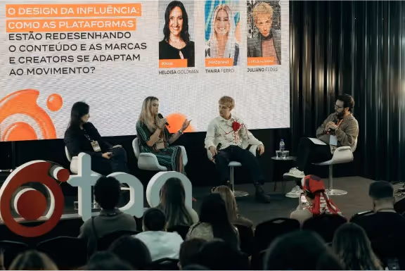 Painel com três palestrantes sentados em cadeiras em um palco, discutindo design da influência diante de uma plateia, com telão exibindo títulos e fotos dos palestrantes.