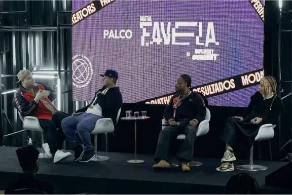 Quatro pessoas sentadas em cadeiras brancas no palco de um painel com fundo roxo e texto 'Palco Digital Favela'.
