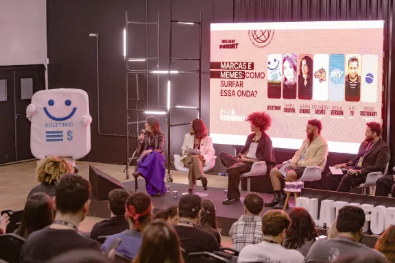 Painel de discussão com cinco pessoas sentadas no palco em frente a uma plateia, com um mascote em formato de aplicativo do Boltehinho à esquerda e uma tela iluminada ao fundo.