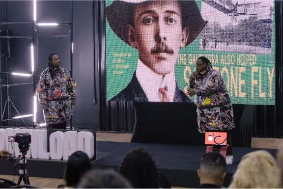 Duas mulheres com roupas coloridas participam de uma apresentação em palco com público, atrás delas tela grande exibindo imagem histórica de um homem com chapéu.