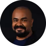 Homem sorridente com barba e cabeça raspada em fundo preto.