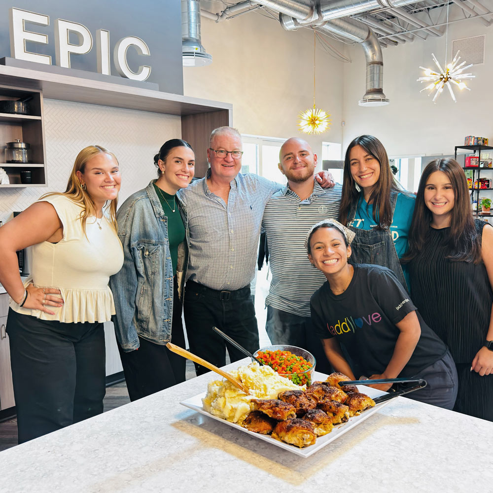 Group of smiling coworkers gathered in a modern kitchen space around a plate of food, beneath a sign that reads “EPIC.”