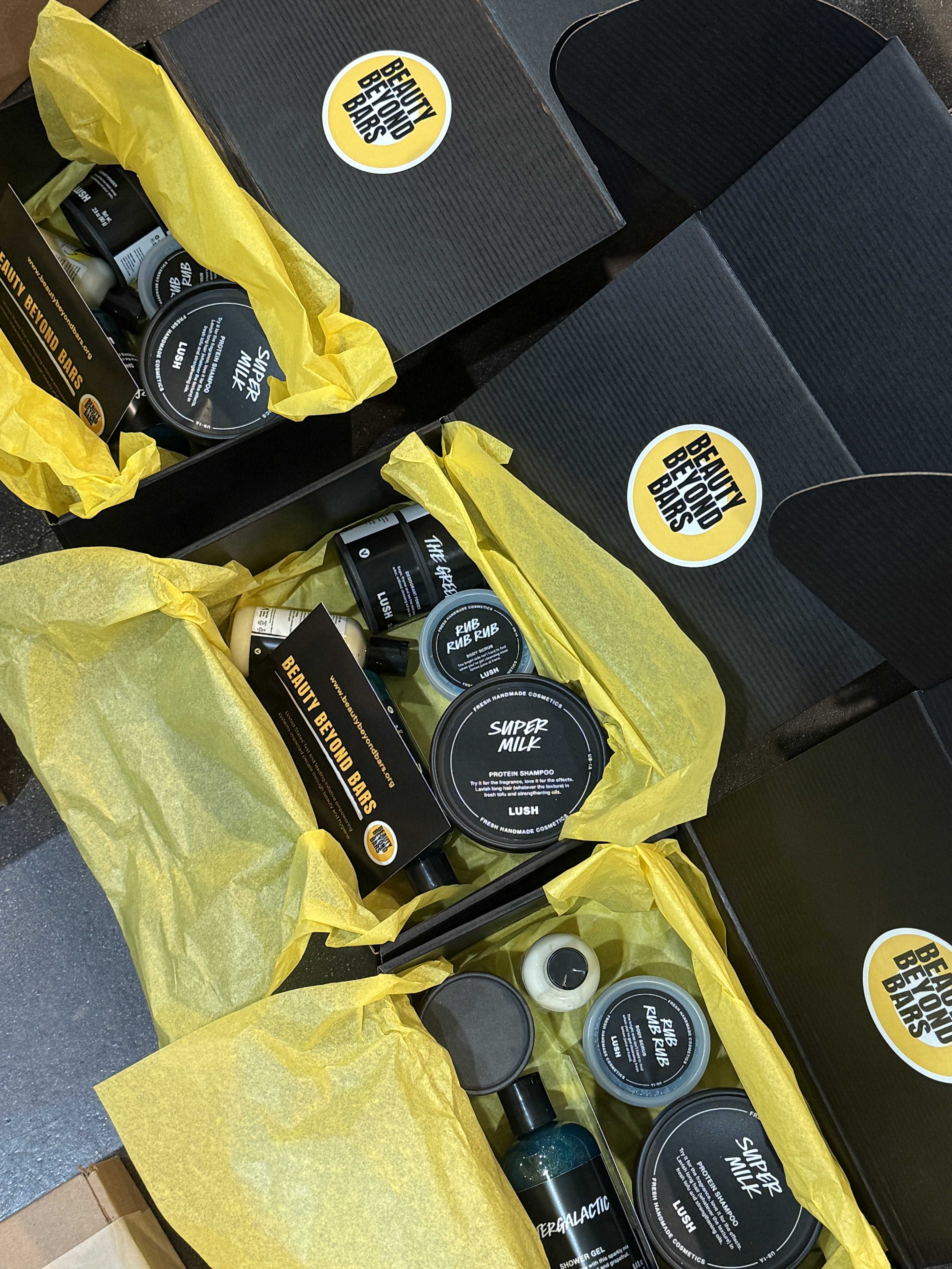Open black gift boxes with yellow tissue paper filled with Lush beauty products including Super Milk protein shampoo and Rub Rub Rub body scrub.