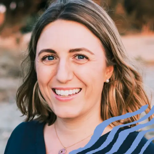 Smiling woman with shoulder-length blonde hair wearing a dark blue top and a necklace, outdoors with blurred background.