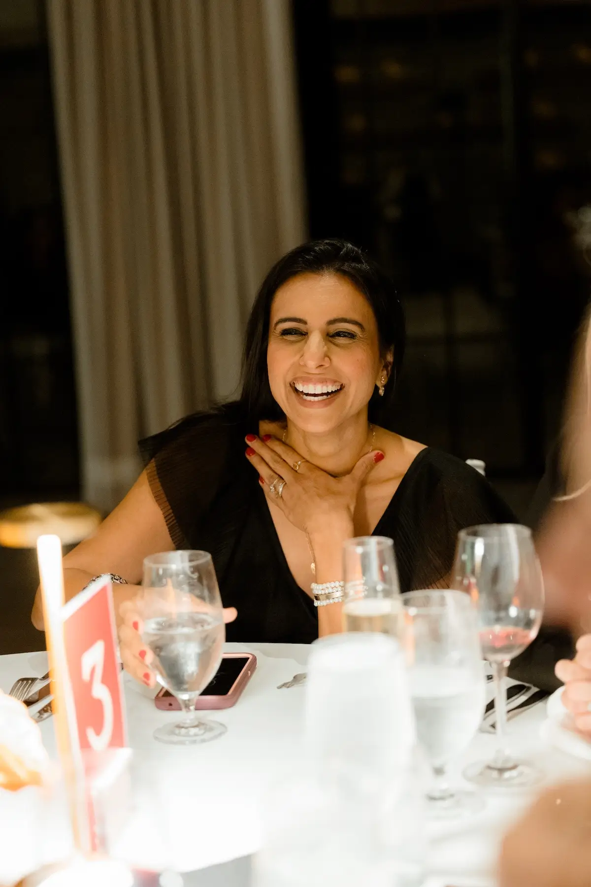 Woman with dark hair laughing and holding a glass of water at a dining table with a red table number 3.
