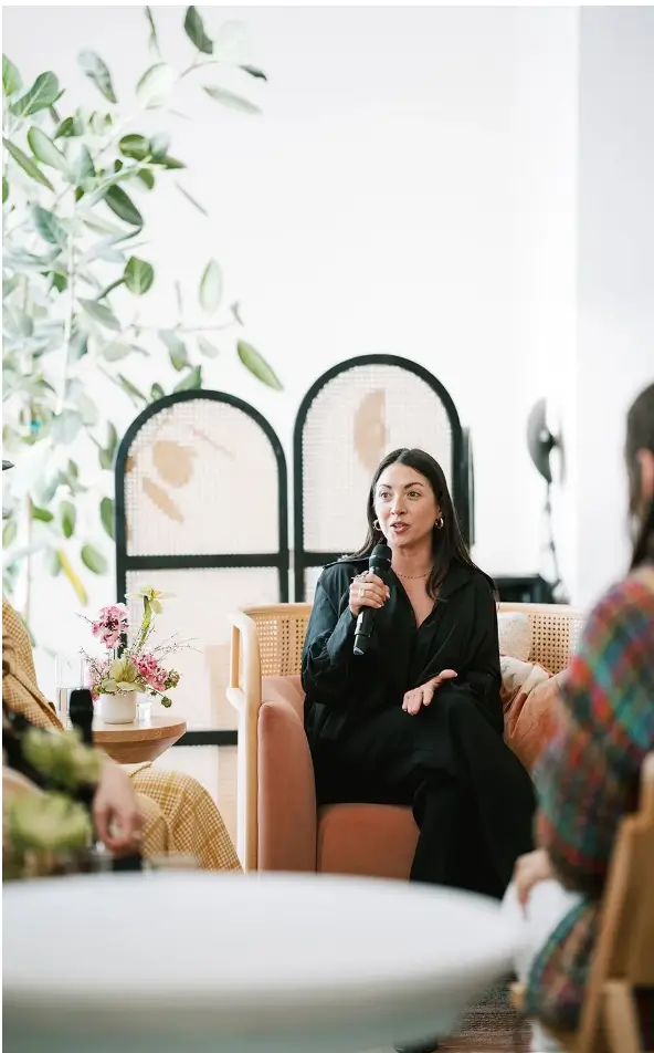 Woman in black outfit sitting on a chair speaking into a microphone during a casual group discussion.