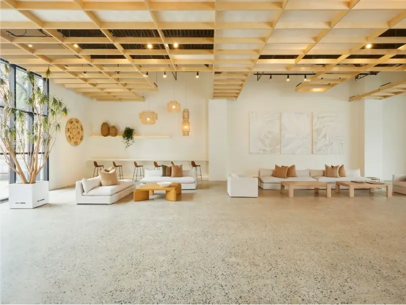 Spacious modern living room with light wooden grid ceiling, large windows, white sofas with brown cushions, wooden coffee tables, and decorative plants.