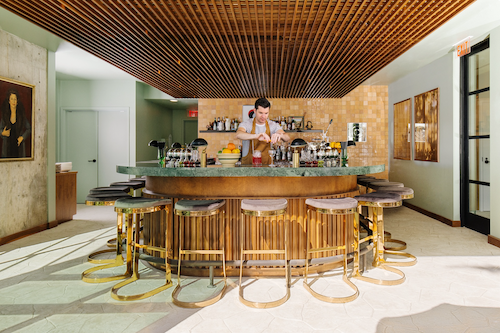 Bartender preparing drinks behind a curved wooden bar with gold stools in a bright, stylish interior.