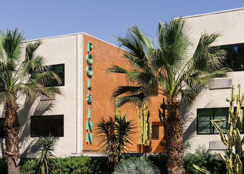 Exterior of a building with palm trees and cactus plants, featuring a vertical sign that reads 'POOL & INN' on an orange brick wall.