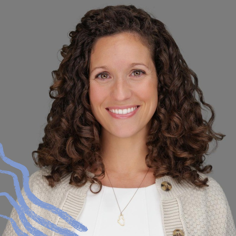 Smiling woman with curly brown hair wearing a beige cardigan and white top against a gray background.