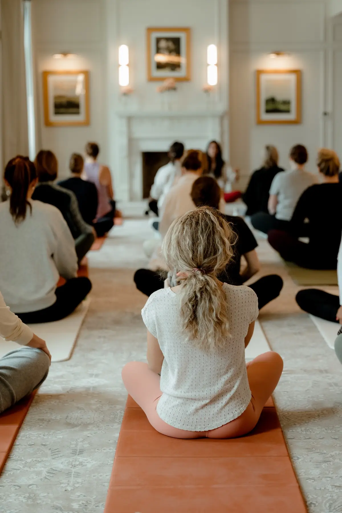 Group of people sitting cross-legged on yoga mats in a room, facing a person leading a meditation session near a fireplace.