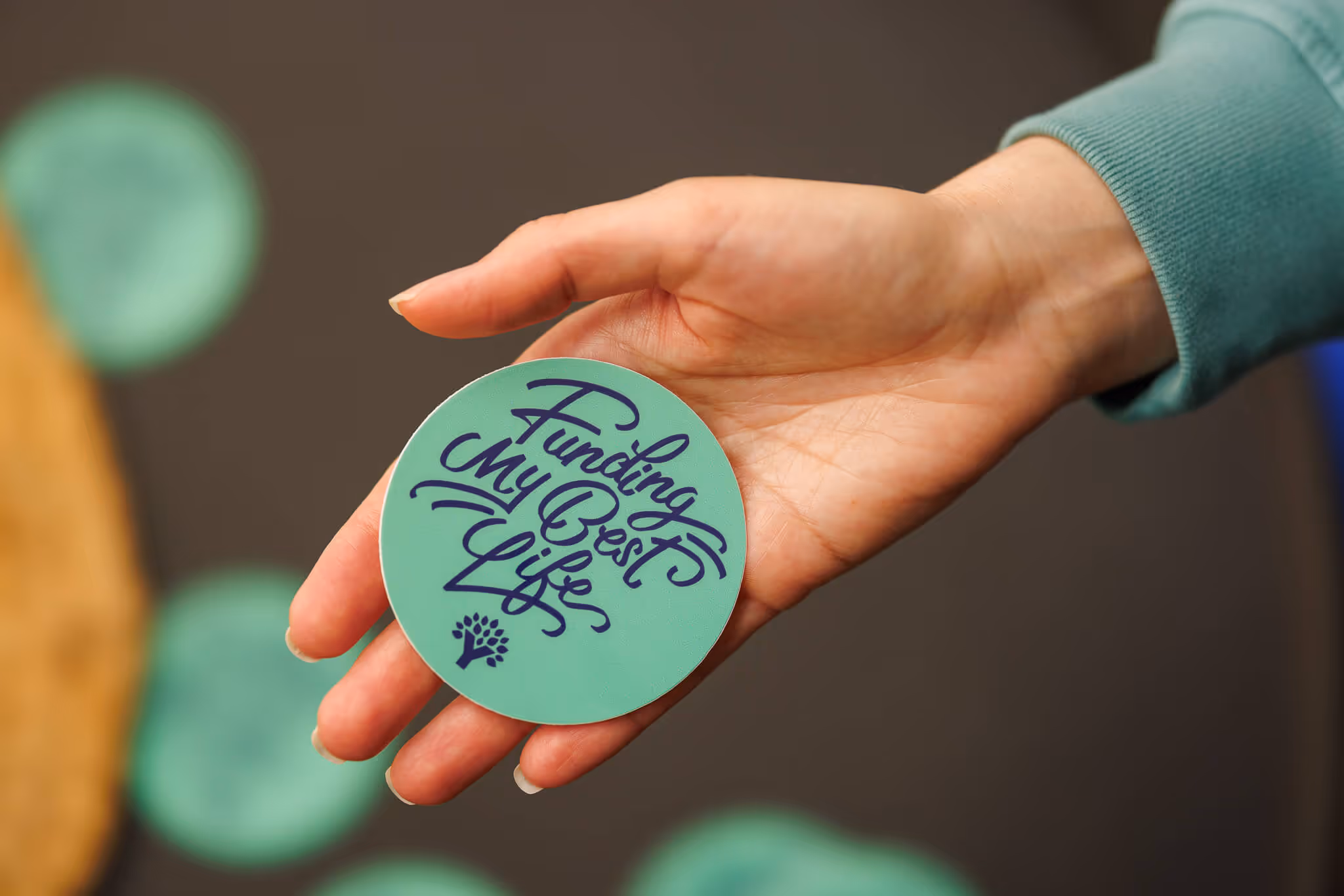 Hand holding a green circular token with the text 'Funding My Best Life' and a small tree icon.
