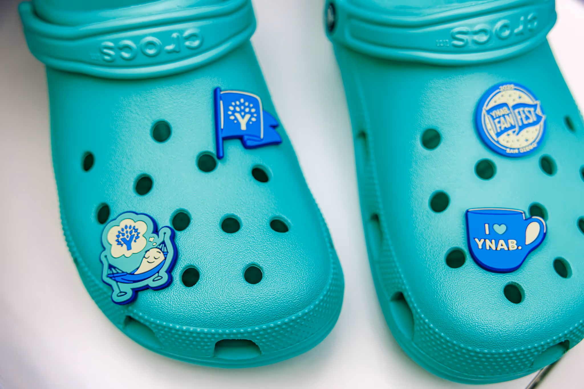 Pair of turquoise Crocs decorated with blue and white pins including a flag, a sleeping character in a hammock, a fan fest badge, and a cup that says 'I love YNAB.'