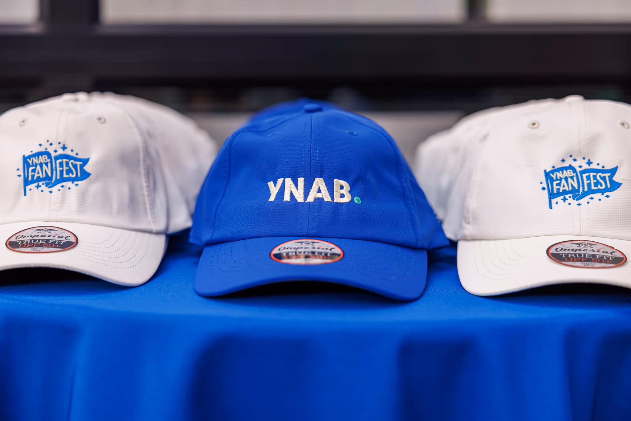 Blue YNAB baseball cap with embroidered logo on blue tablecloth, beside white caps.