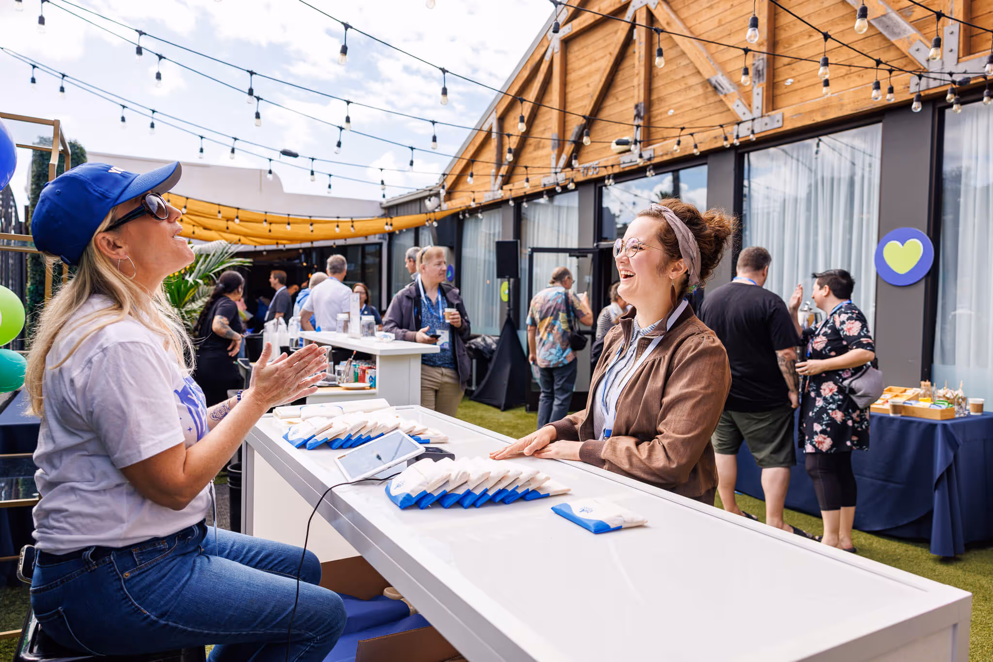 Two people smiling and chatting at the YNAB Fan Fest merch table, with others mingling in the background under string lights.