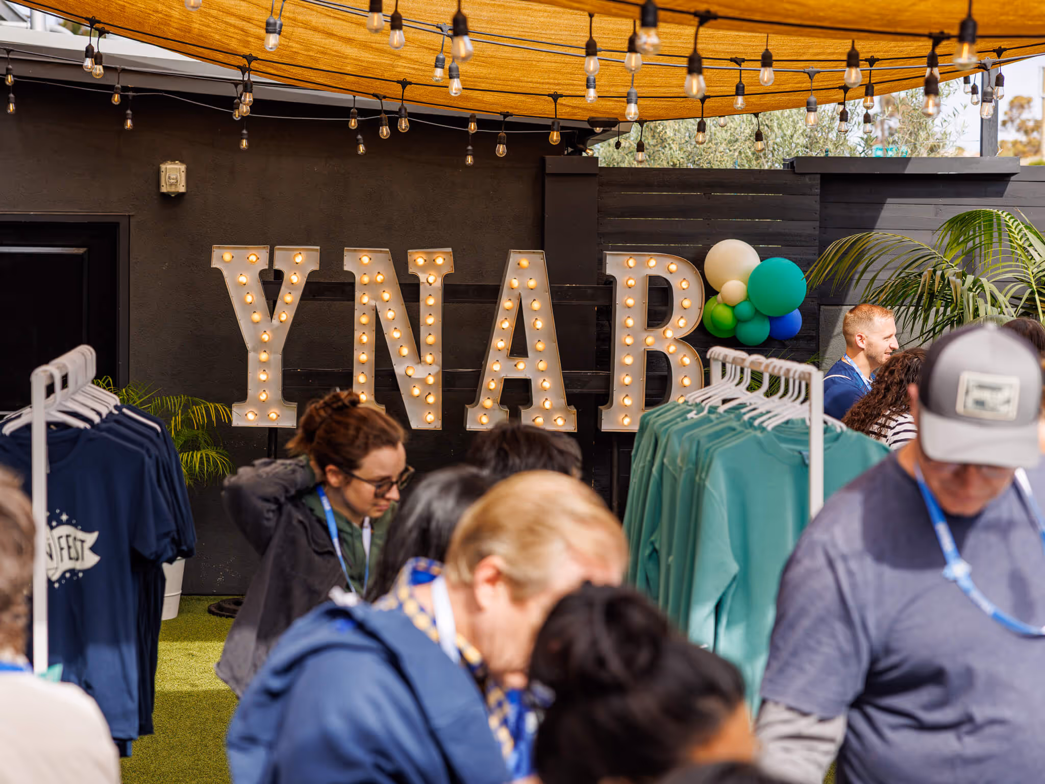 Crowd browsing YNAB merchandise racks at Fan Fest, with large illuminated YNAB letters and colorful balloon decor in the background.