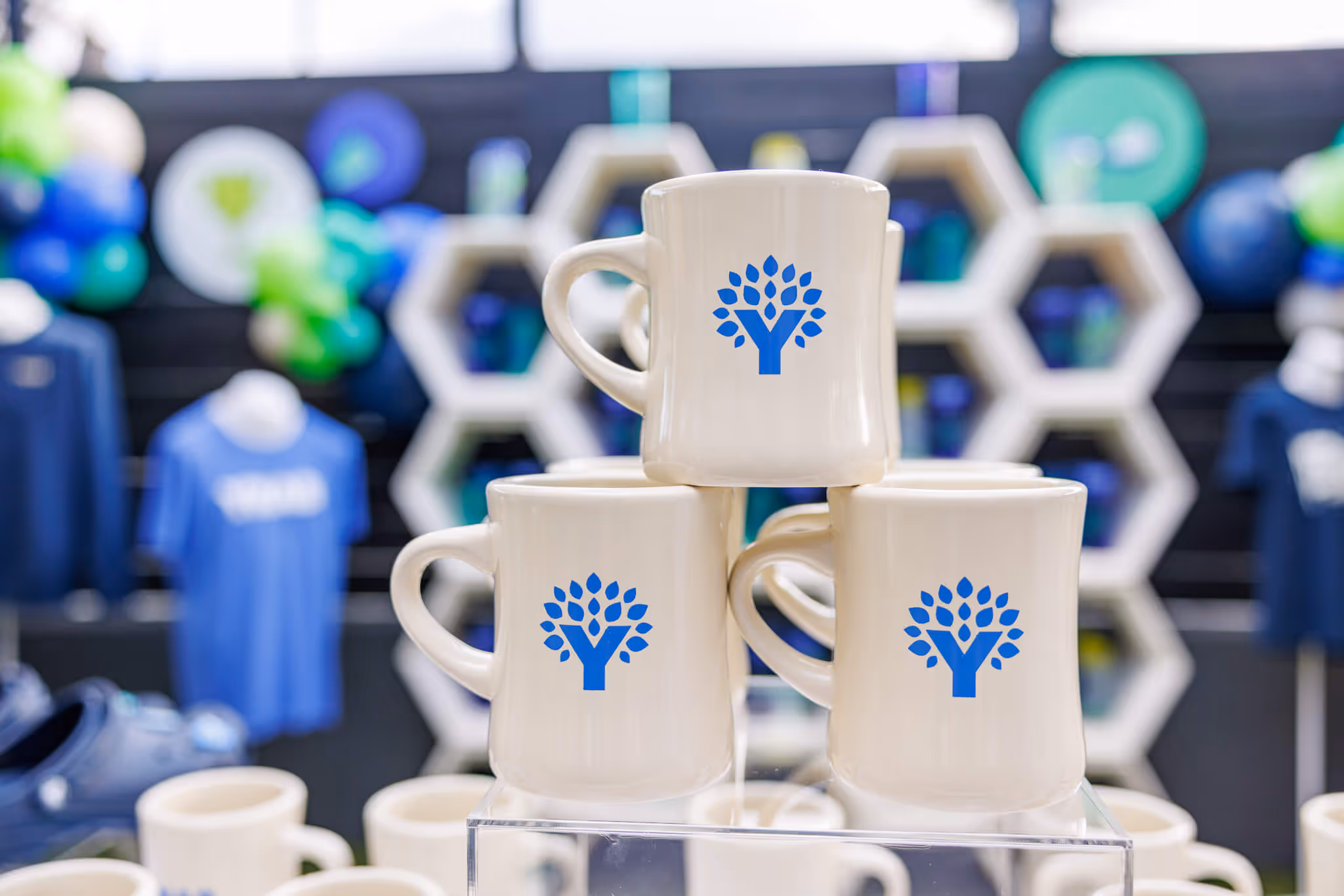 Cream-colored mugs with blue YNAB tree logos displayed on a clear stand in the Fan Fest merch area, with other branded products in the background.