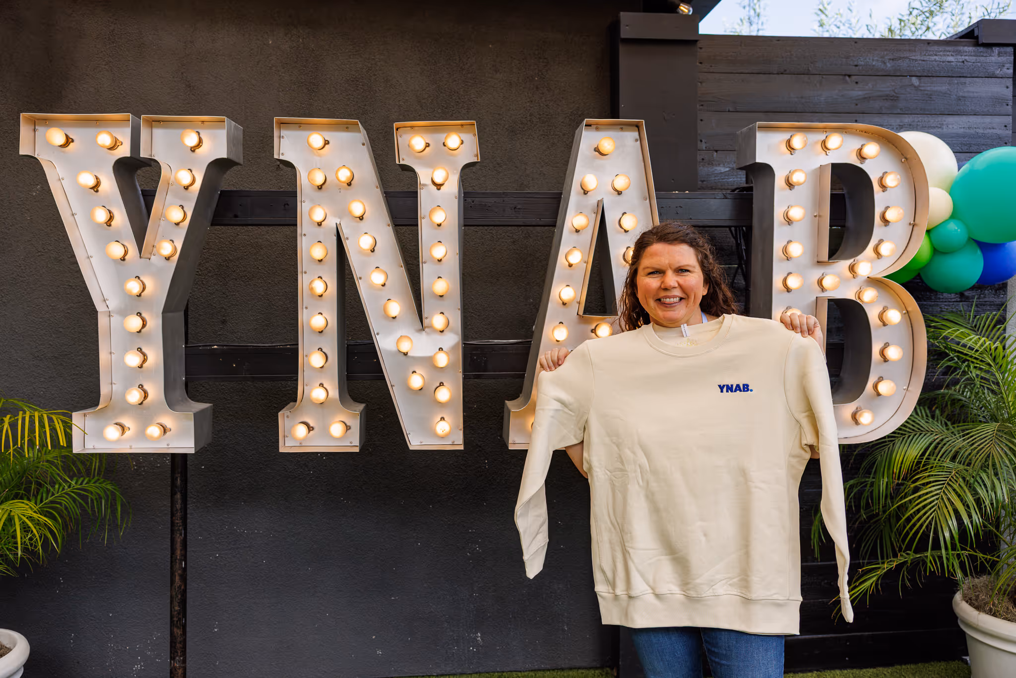 Smiling woman holding up a cream-colored sweatshirt with a small blue YNAB logo in front of large illuminated letters spelling YNAB on a black background with plants and balloons.