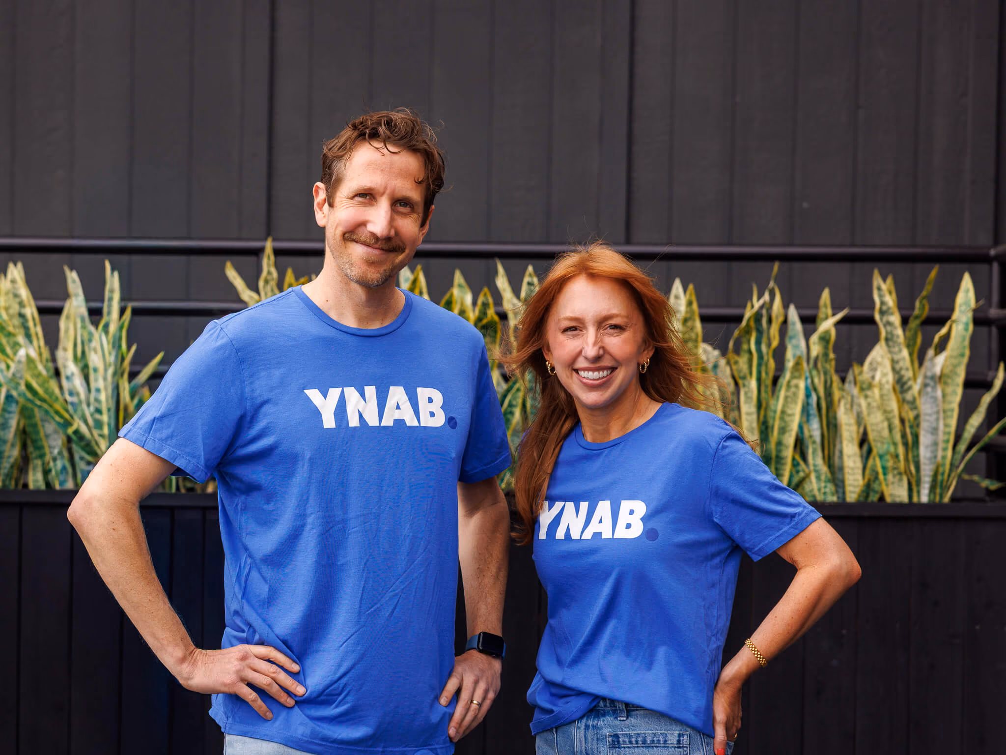 A man and a woman stand side by side smiling, both wearing blue t-shirts with the text 'YNAB' printed in white, posed in front of tall green plants and a dark wall.