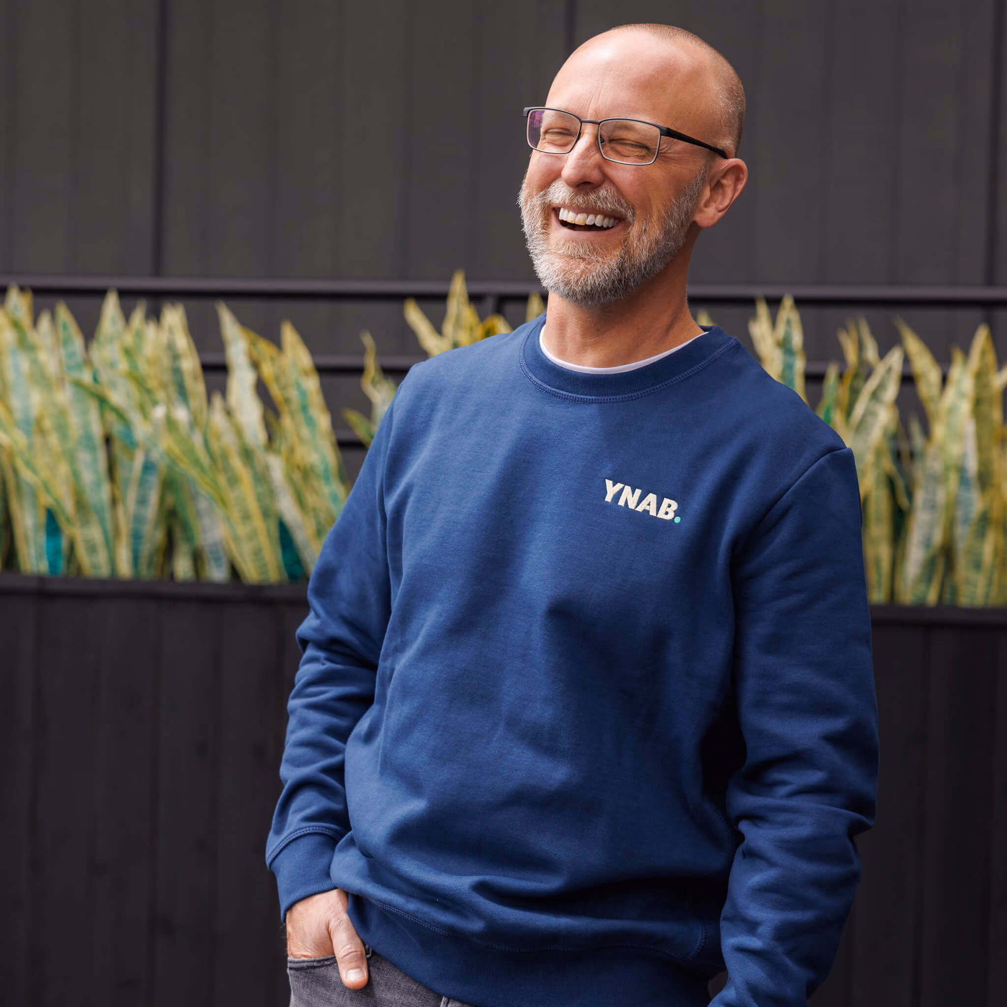 Smiling man with glasses and a beard wearing a blue YNAB sweatshirt standing in front of a dark fence with green leafy plants.